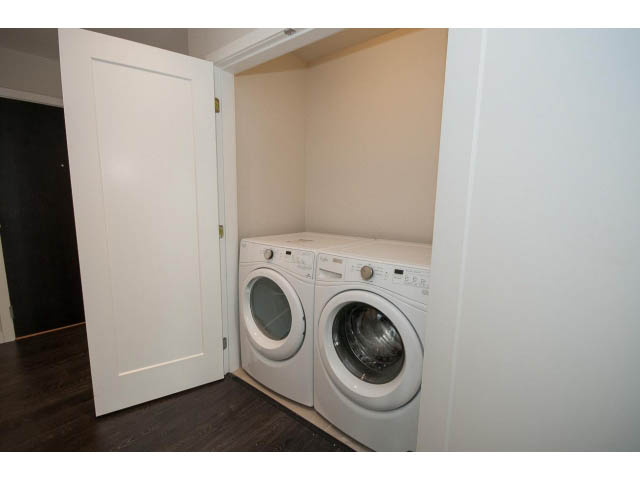 Laundry room with white washer and dryer, hardwood floors, and a door leading to another room. - 303 - 3825 Cate's Landing, North Vancouver