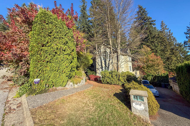 Front of the house among red and green trees and shrubs, a sloped lawn and the stone 3858 address post by the driveway.