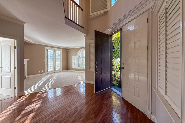 Two-storey foyer with hardwood floors and double front doors standing open, looking into the carpeted living room with French