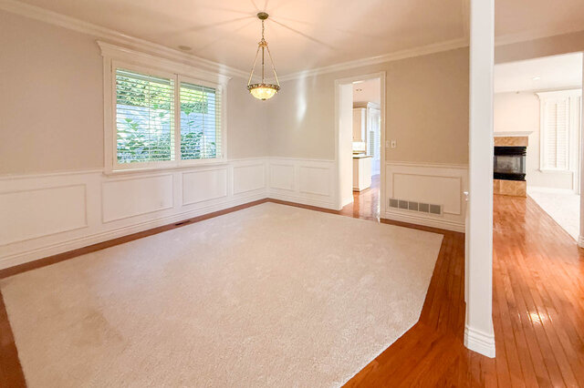 Separate dining room with white wainscoting, a pendant light, carpet bordered by hardwood floors and a window onto greenery, 