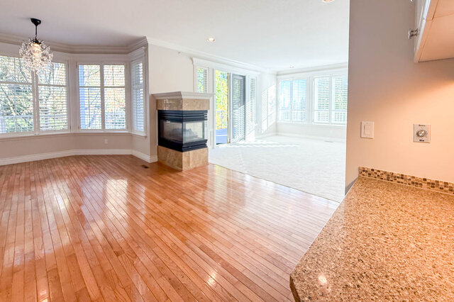 Open family room past a granite counter: hardwood floors, a three-sided gas fireplace in a marble surround, a bay of shuttere