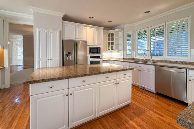 Large granite island with white cabinets, a stainless fridge, built-in microwave and wall oven, and a sink under shuttered wi