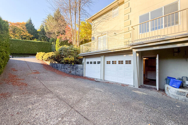 Side of the house with the double garage doors off a long driveway, a balcony above and a stone retaining wall. - 3858 Sauve Place, North Vancouver