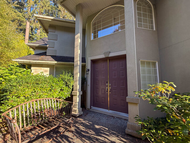 Covered front entry with tall double doors, an arched window above and rhododendrons beside a garden bench. - 3858 Sauve Place, North Vancouver