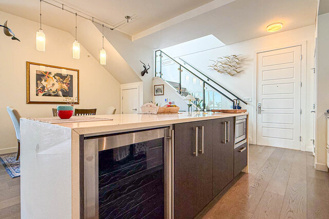 Furnished kitchen with quartz island, dark wood cabinetry, and hardwood floors beneath a glass-railed staircase.