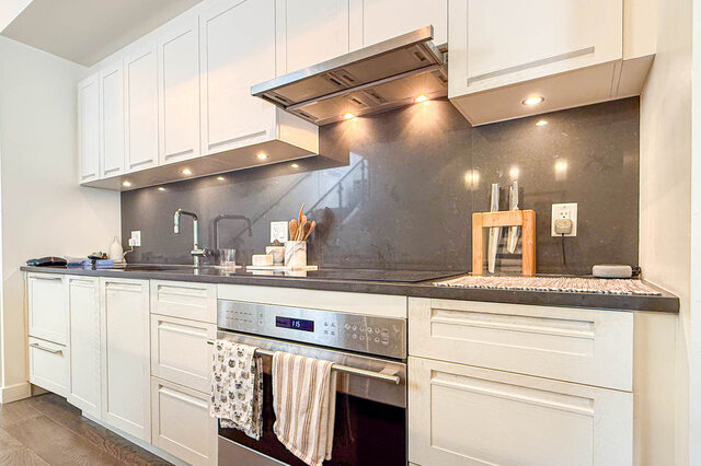 Modern kitchen with white shaker cabinets, dark granite countertops, and a stainless steel range hood. - 503-105 West 2nd Street, North Vancouver
