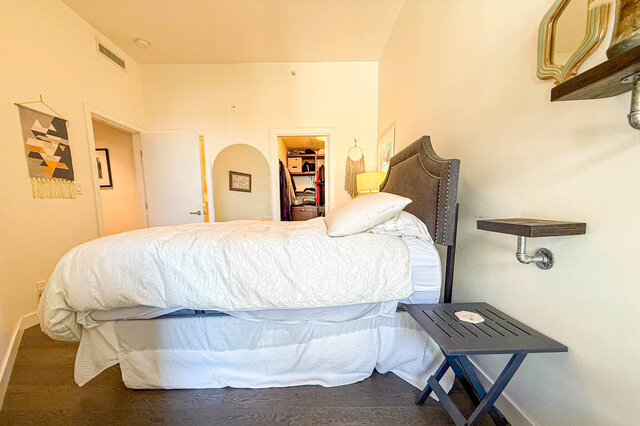 Bedroom with white bedding, gray upholstered headboard, dark hardwood floors, and geometric macrame wall art.