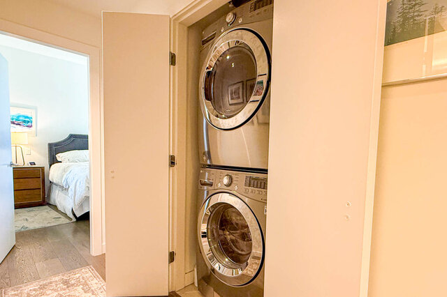 Stacked washer and dryer in a built-in closet, with hardwood floors and a bedroom visible through the open doorway.