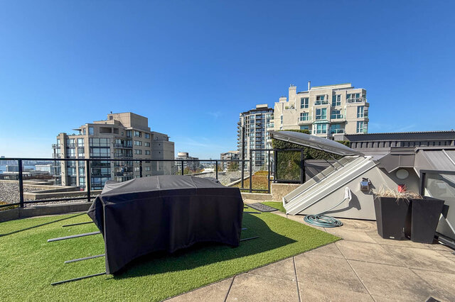 Rooftop terrace with artificial turf, a covered grill, and a clear view of surrounding high-rise condos under a blue sky.