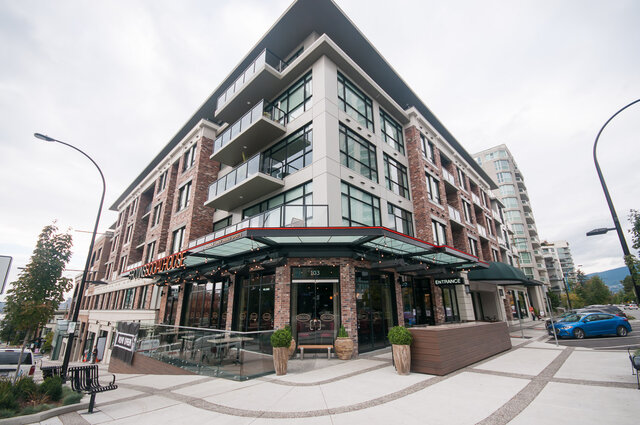 Modern corner condo building with brick facade, glass balconies, and a ground-floor cafe under a glass canopy.