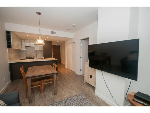 Bright kitchen with dark countertops and hardwood floors, open to a cozy living area with a large flat-screen TV.
