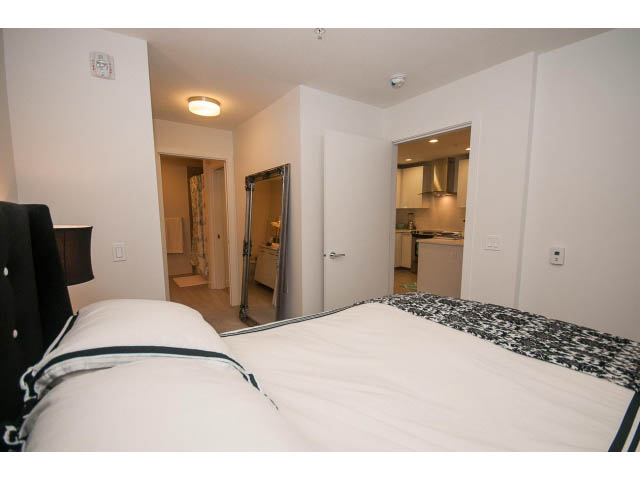 Bright bedroom with white bedding and black accents, leading to a modern kitchen with light countertops and hardwood floors.