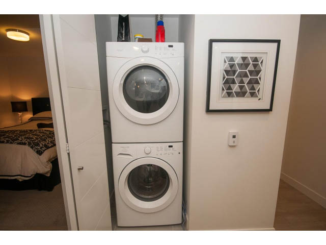 Bright laundry room with stacked washer and dryer, white walls, and geometric art. - 416 - 733 West 3rd St, North Vancouver