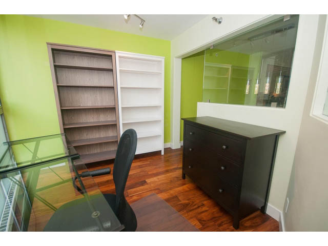 Bright and modern den with hardwood floors, green walls, and a glass partition, featuring a desk and bookshelf.