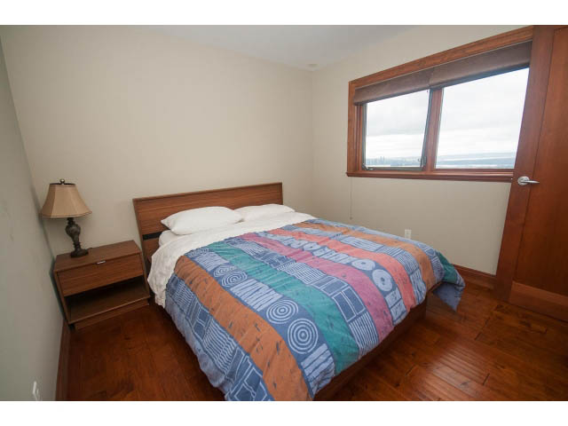 Bright bedroom with hardwood floors and a colorful bedspread, overlooking a scenic view. - Upper - 4407 Prospect Rd, North Vancouver