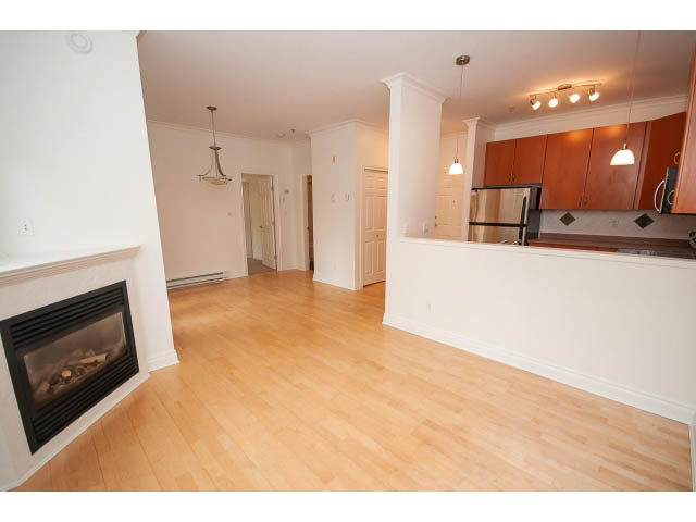 Bright kitchen with hardwood floors and white cabinetry, open to a cozy living area with a fireplace. - 104 - 128 West 21st St, North Vancouver
