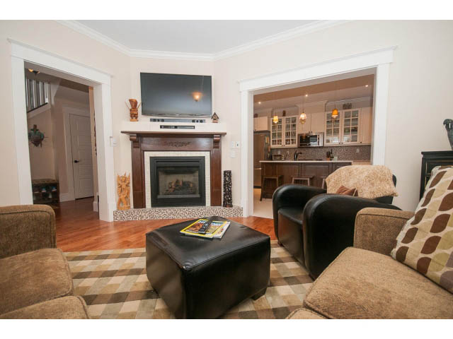 Furnished living room with hardwood floors, checkered rug, and kitchen in background. - 266 W 6th St, North Vancouver