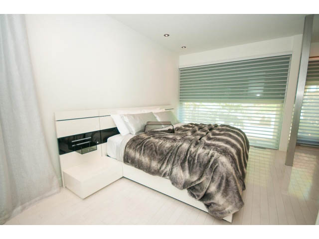 Spacious bedroom with modern white furniture, hardwood floors, and a large window with blinds. - 5517 Ocean Place, West Vancouver