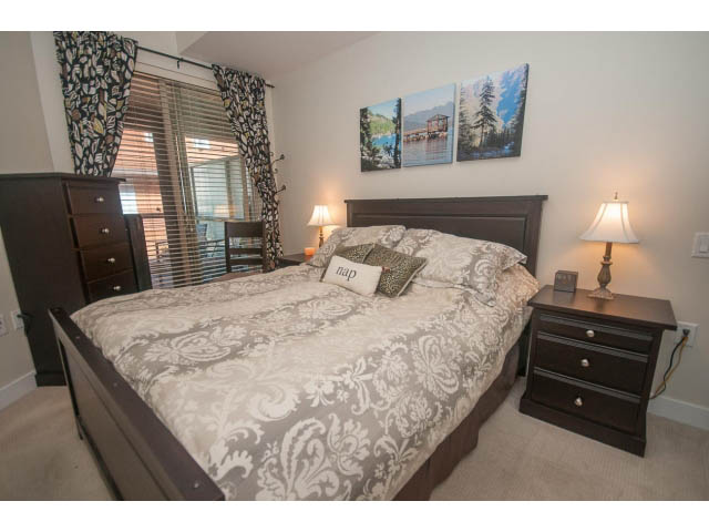 Furnished 2-bedroom condo with a cozy bed, patterned bedding, and a nightstand with a lamp. - 205 - 116 West 23rd, North Vancouver