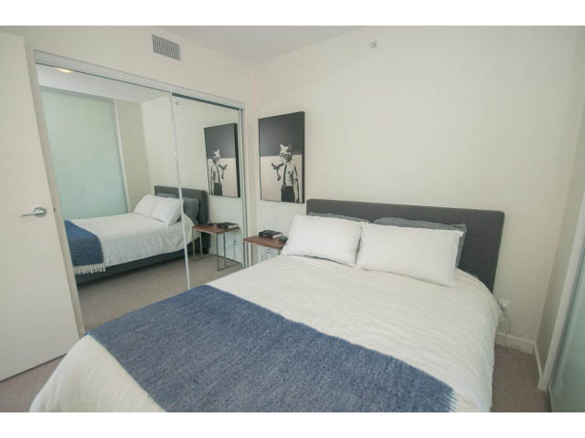 Furnished 1-bedroom + den with two queen beds, modern decor, and a sliding glass door leading to a private balcony.