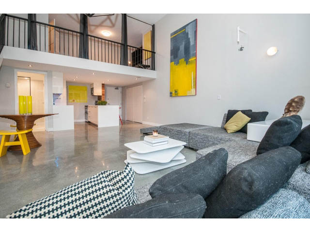 Modern open-concept living space with concrete floors, contemporary furniture, and a vibrant yellow accent wall.