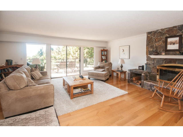 Spacious living room with hardwood floors, stone fireplace, and large windows offering a view. - 3855 Bayridge Ave, West Vancouver