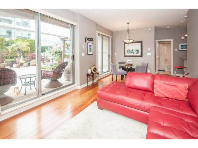 Furnished living room with red leather couch and hardwood floors, leading to a balcony with city view. - 310 - 160 E 13th St, North Vancouver