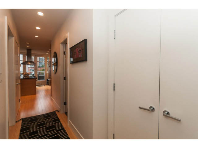 Bright hallway with hardwood floors and modern door handles, leading to a kitchen with stainless steel appliances.