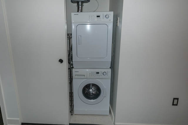 Bright laundry room with stacked washer and dryer, white walls, and visible plumbing. - 1001 - 175 West 2nd Street, North Vancouver