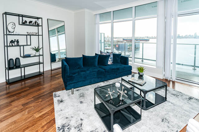 Bright living room with hardwood floors, a blue sectional sofa, and a city view. - 702 - 185 Victory Ship Way, North Vancouver