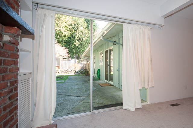 Living room with sliding glass door leading to a patio, brick fireplace, and light-colored curtains. - 4037 Ripple Road, West Vancouver