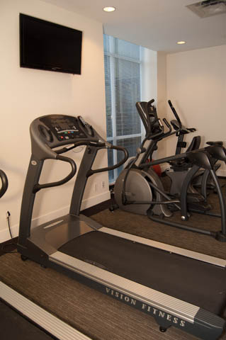 Bright gym with a treadmill and elliptical, featuring a TV mounted on the wall and a window with blinds. - 1607 - 888 Homer St, Vancouver Downtown