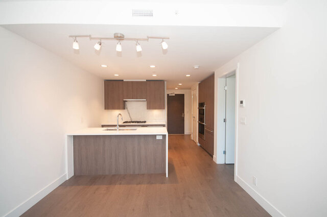 The kitchen features a central island with a sink, wooden cabinets, and track lighting on the ceiling. - 203 -2785 Library Lane, North Vancouver