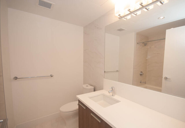 This is a bathroom with a white countertop sink, toilet, mirror, and towel bar. - 203 -2785 Library Lane, North Vancouver