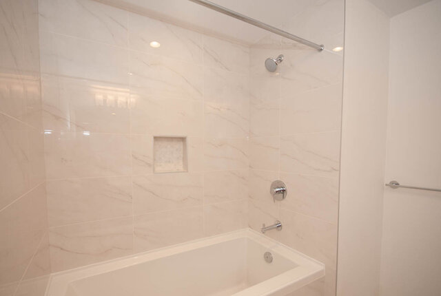 This is a bathroom with a white bathtub, marble tile walls, a showerhead, and shower controls. - 203 -2785 Library Lane, North Vancouver