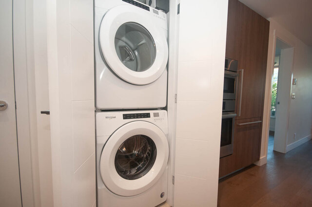 The laundry room contains a stacked washer and dryer, tiled walls, and hardwood floors. - 203 -2785 Library Lane, North Vancouver