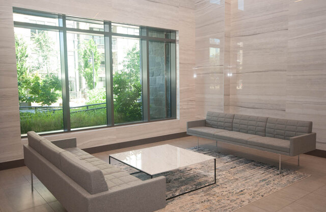The lobby features two sofas, a coffee table, an area rug, and floor-to-ceiling windows overlooking greenery.