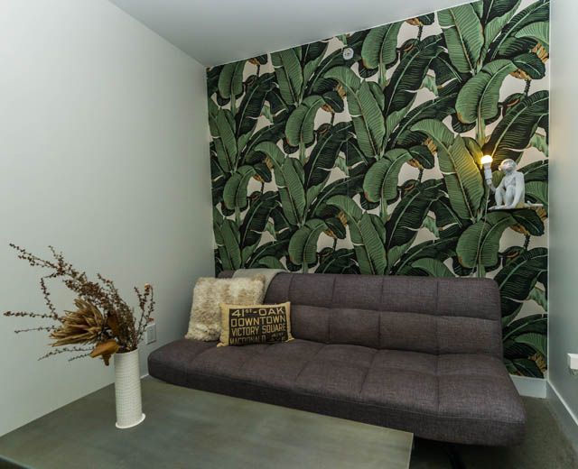 Furnished living room with banana leaf wallpaper, dark sofa, and decorative pillows. - 501 - 231 E Pender Street, Vancouver Downtown