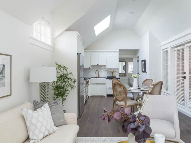 Bright living room with hardwood floors and white furniture, leading to a kitchen with stainless steel appliances.