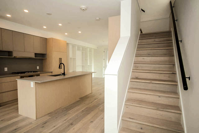 Modern kitchen with light wood cabinets and island, leading to a spacious living area with hardwood floors and a staircase.