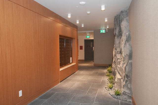 Spacious hallway with wood paneling and rock accent wall, leading to a door and emergency exit sign. - 202-6699 Nelson Avenue, West Vancouver