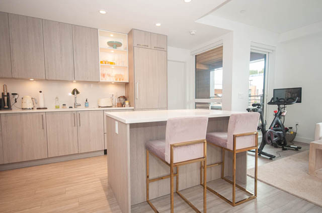 Furnished kitchen with light wood cabinets and hardwood floors, open to a living area with a city view. - 211 - 733 E 3rd St, North Vancouver