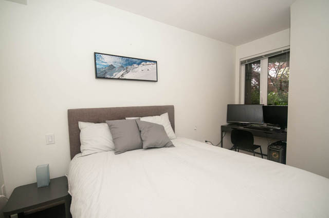 Furnished 1-bedroom + den with a cozy bed, modern artwork, and a sliding glass door leading to a private patio. - 211 - 733 West 14th, North Vancouver