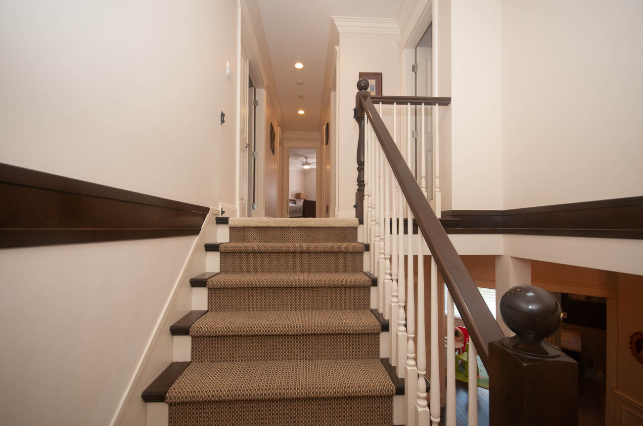 Furnished hallway with carpeted stairs leading to upper floor, white walls, dark trim, and wooden railings.