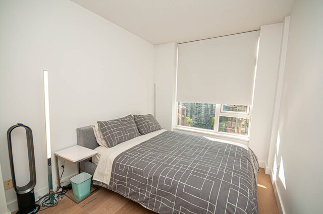 A bedroom with a bed, side table, window showing city buildings, hardwood floors, and a floor lamp. - 1306-480 Robson Street, Vancouver Downtown