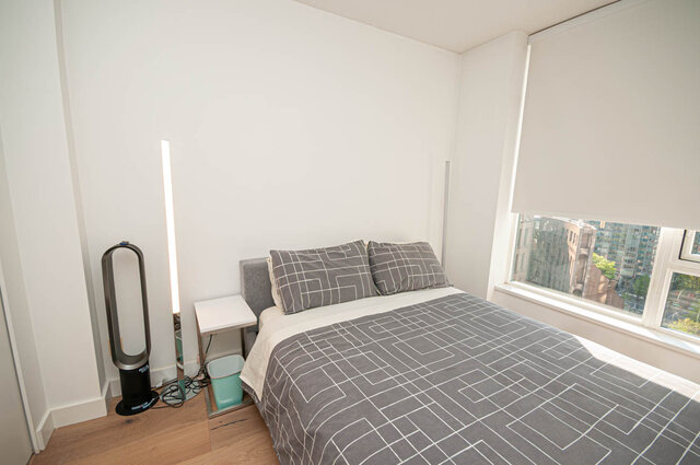 A bedroom with a bed, small nightstand, hardwood floors, and a window showing city buildings. - 1306-480 Robson Street, Vancouver Downtown