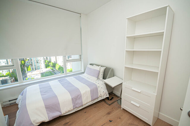 This bedroom contains a bed with striped bedding, a small table, a tall bookshelf, and large windows with a city view.