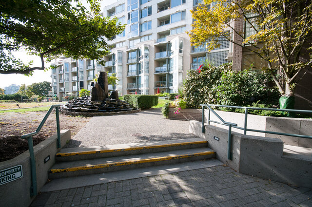 Building exterior featuring concrete steps with railings, greenery, and a multi-story residential building in the background.