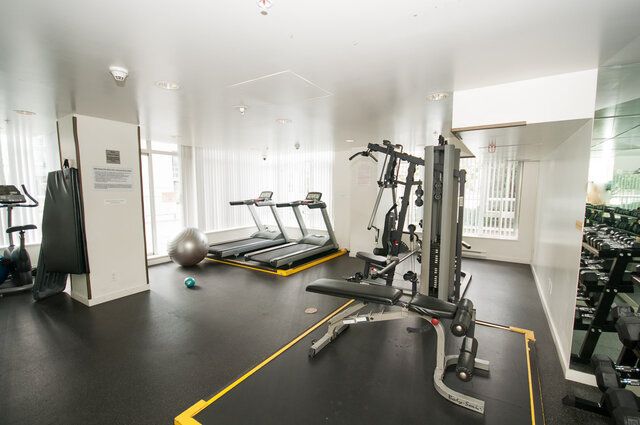 The gym contains treadmills, weight machines, dumbbells, and a stability ball on a black floor with yellow markings.