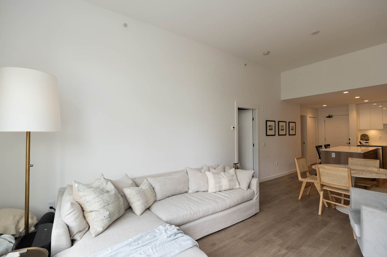 Bright living room with white couch, hardwood floors, and kitchen counter in background. - 308 - 715 W15th Street, North Vancouver